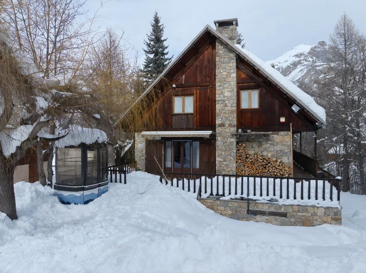 Chalet Pegulu sous la neige, façade en mélèze et cheminée en pierre, montagnes en arrière-plan